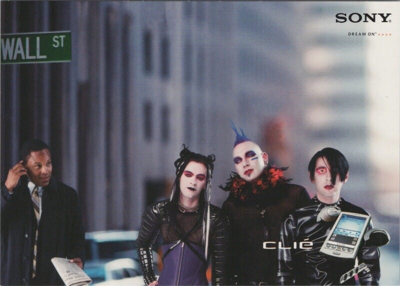 Wall Street. A businessperson on their cellphone looks incredulous at three people composited into the scene who are done up in mallgoth style: white pancake foundation, heavy eye makeup, pleather bondage gear, cyberfalls, a mohawk, etc. A Sony Clié with mp3 attachment is superimposed in the corner over one of them.