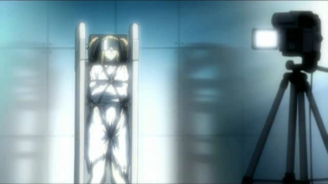 Misa bound upright, face covered, with bondage straps around her body and a video camera pointed at her 